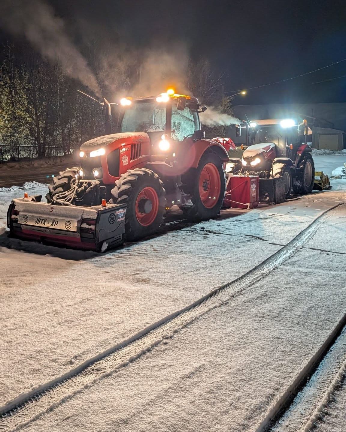 Deux tracteurs attelés de chasse-neige, phares et gyrophares allumés, arrêtés sur une rue enneigée la nuit